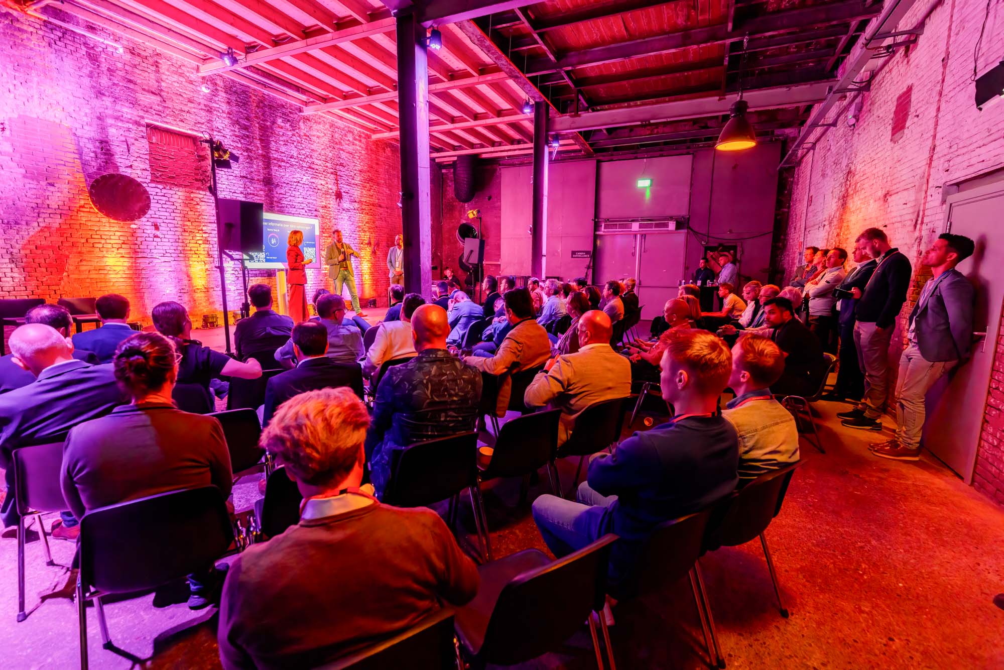 Two men in white shirts holding large arrow signs labeled 'HOPE' and 'NaaS' inside a spacious, industrial-style event space with pink and purple lighting and groups of people socializing.