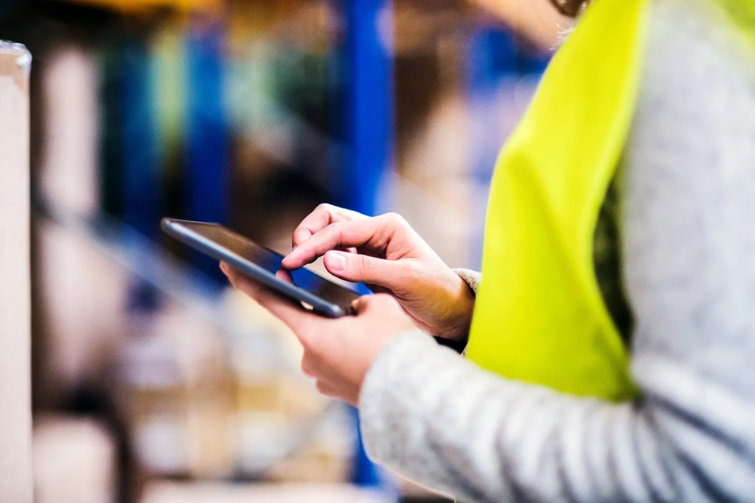 A close up of a worker wearing a yellow safety vest while typing on a tablet in a warehouse.
