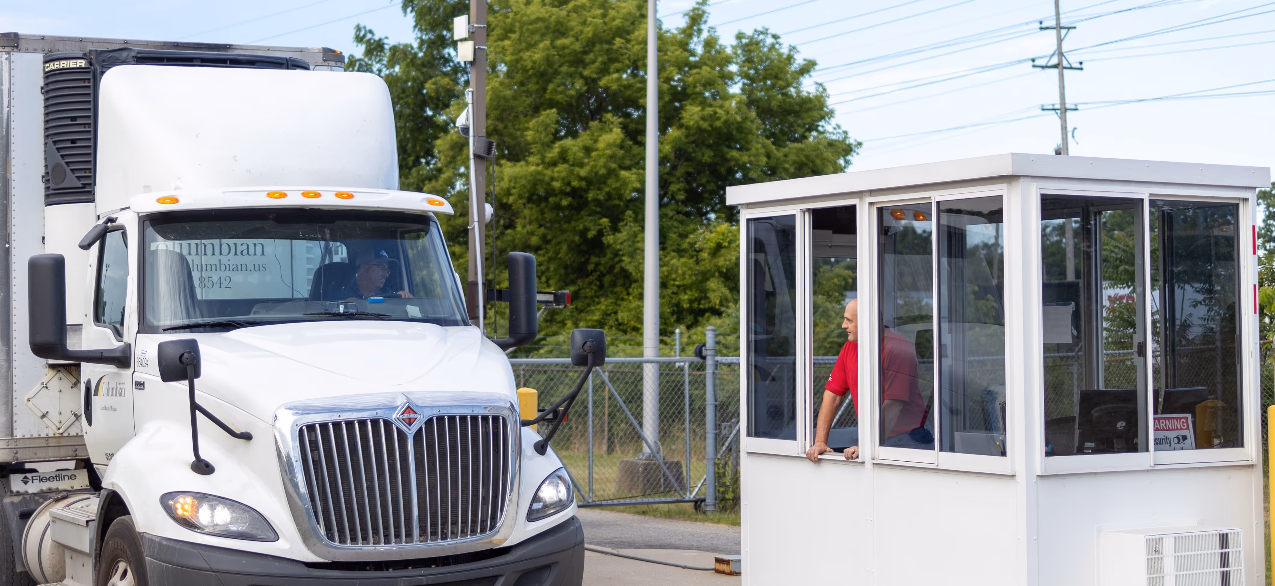 A truck stopping at a gate with a guard greeting the driver through the window.