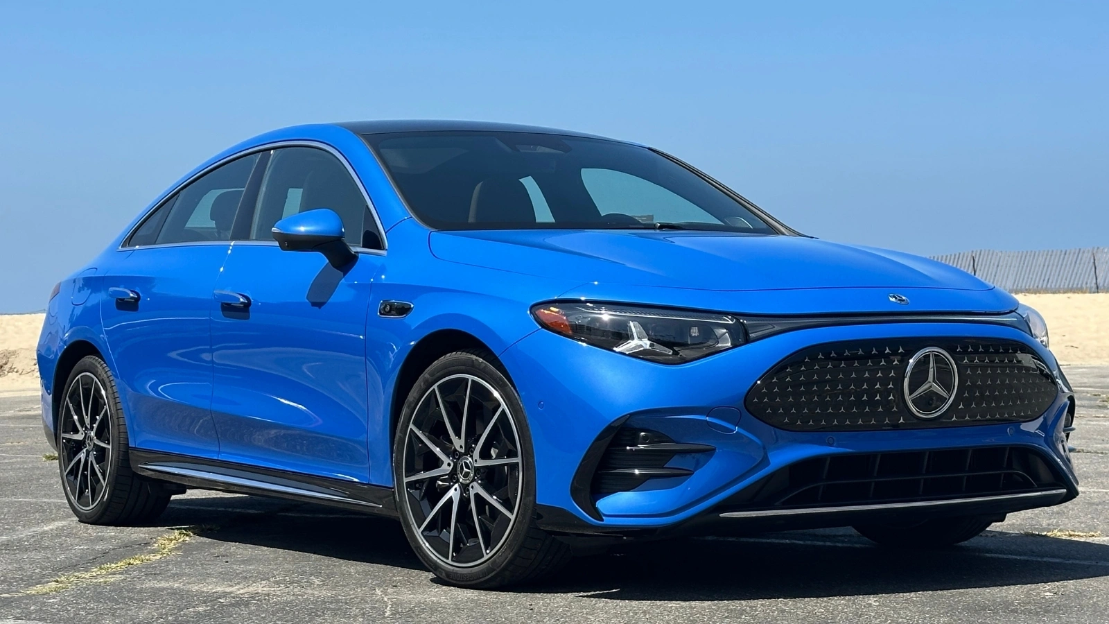 A blue 2027 Mercedes-Benz CLA 250+ Electric angled on pavement in front of a beach and blue skies