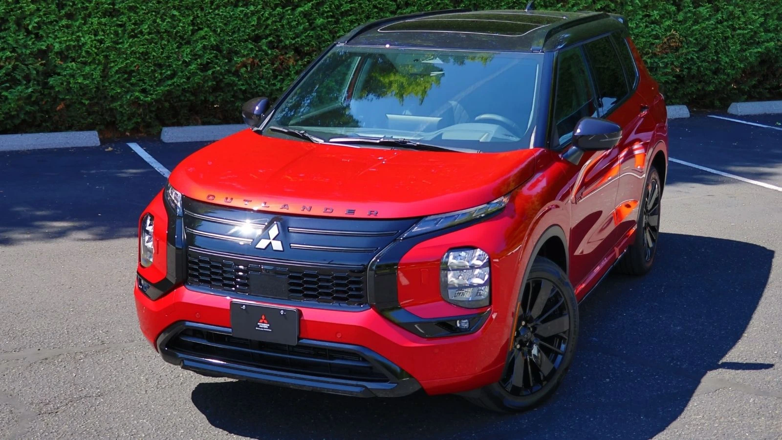 Side profile of a red 2026 Mitsubishi Outlander PHEV in front of a large green hedge
