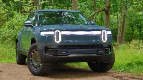Imagen frontal en ángulo de una Rivian R1T 2026 azul en un camino de tierra frente a una zona boscosa 