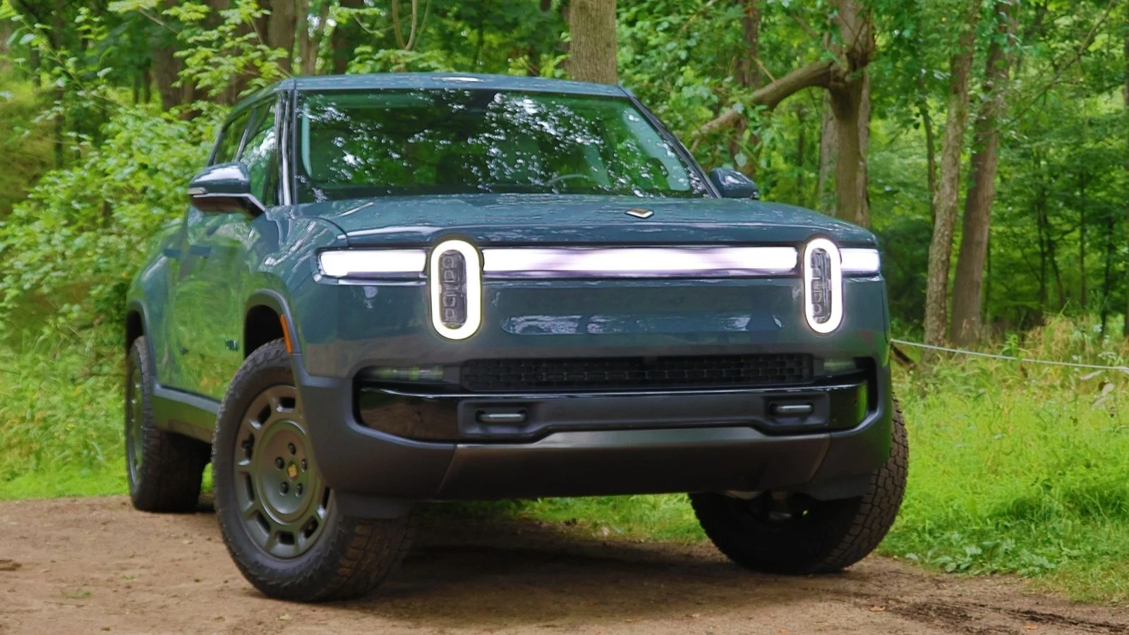 Imagen frontal en ángulo de una Rivian R1T 2026 azul en un camino de tierra frente a una zona boscosa