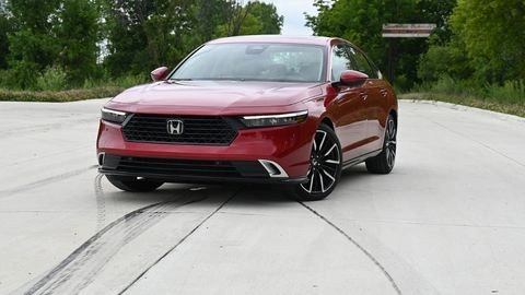 Red 2026 Honda Accord Hybrid sitting on an angle on a concrete pad surrounded by trees.