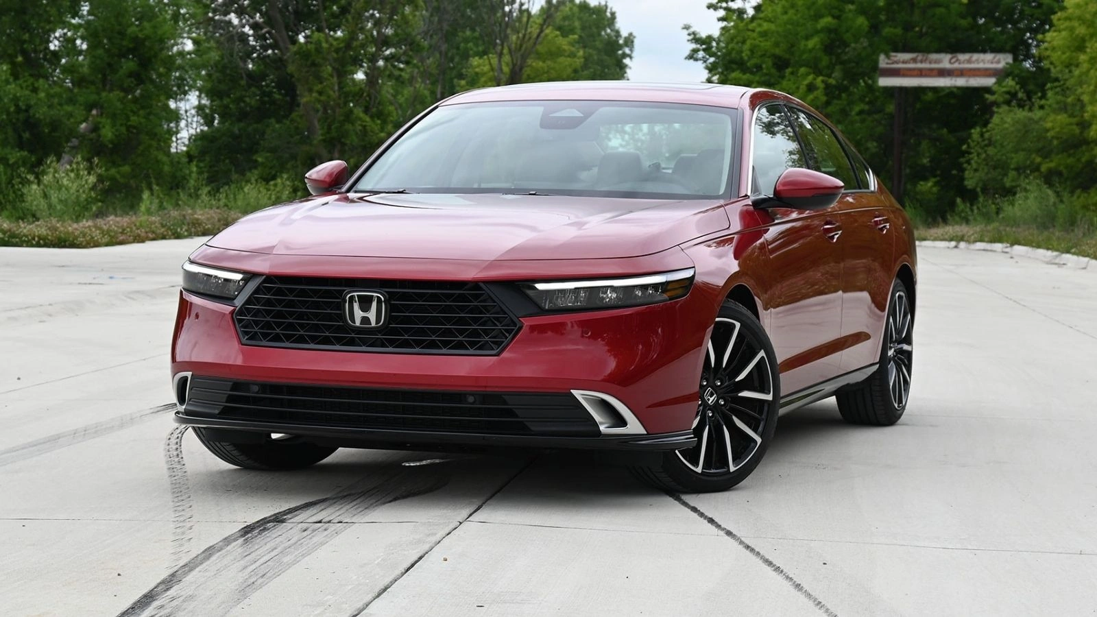 Red 2026 Honda Accord Hybrid sitting on an angle on a concrete pad surrounded by trees.
