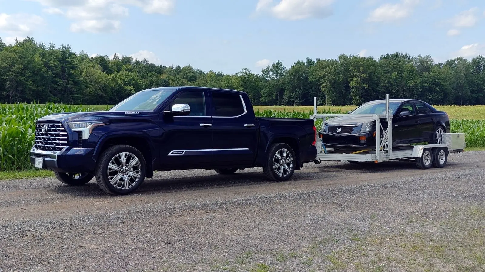 Toyota Tundra Capstone Hybrid 2026 azul remolcando un automóvil Cadillac negro frente a un campo de maíz