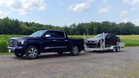 Blue 2026 Toyota Tundra Capstone Hybrid towing a black Cadillac car in front of a corn field