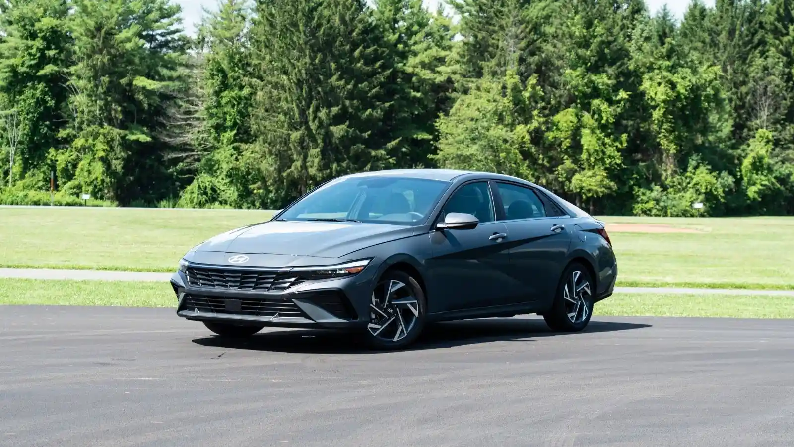 2026 Hyundai Elantra Hybrid parked in an empty parking lot on an angle in front of green grass and a heavily forested area