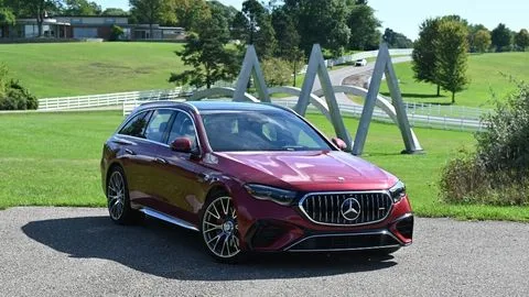 Un Mercedes-AMG E 53 Hybrid Wagon rojo de 2026 aparcado en ángulo frente a una escultura y una propiedad ajardinada