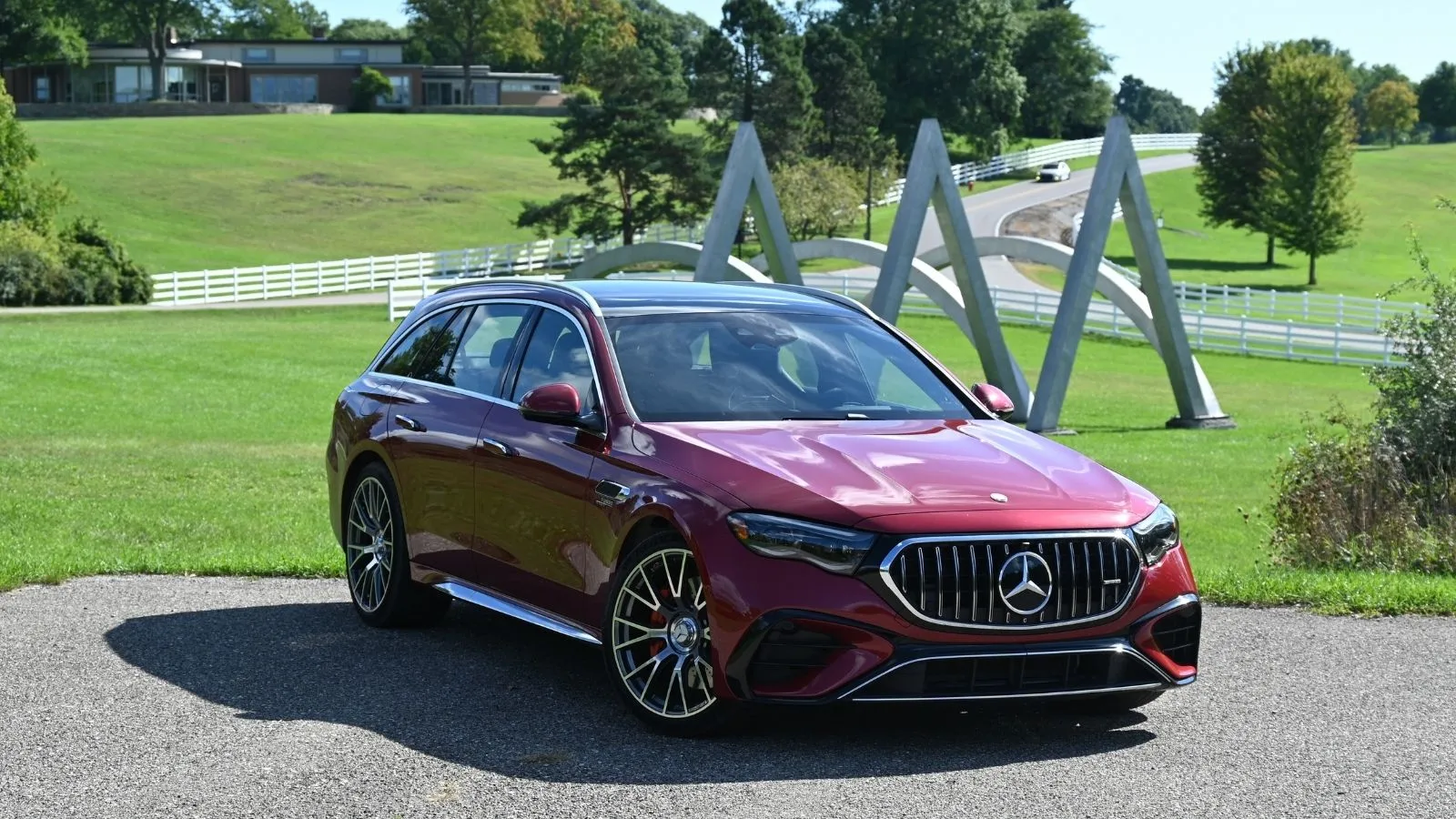 Un Mercedes-AMG E 53 Hybrid Wagon rojo de 2026 aparcado en ángulo frente a una escultura y una propiedad ajardinada