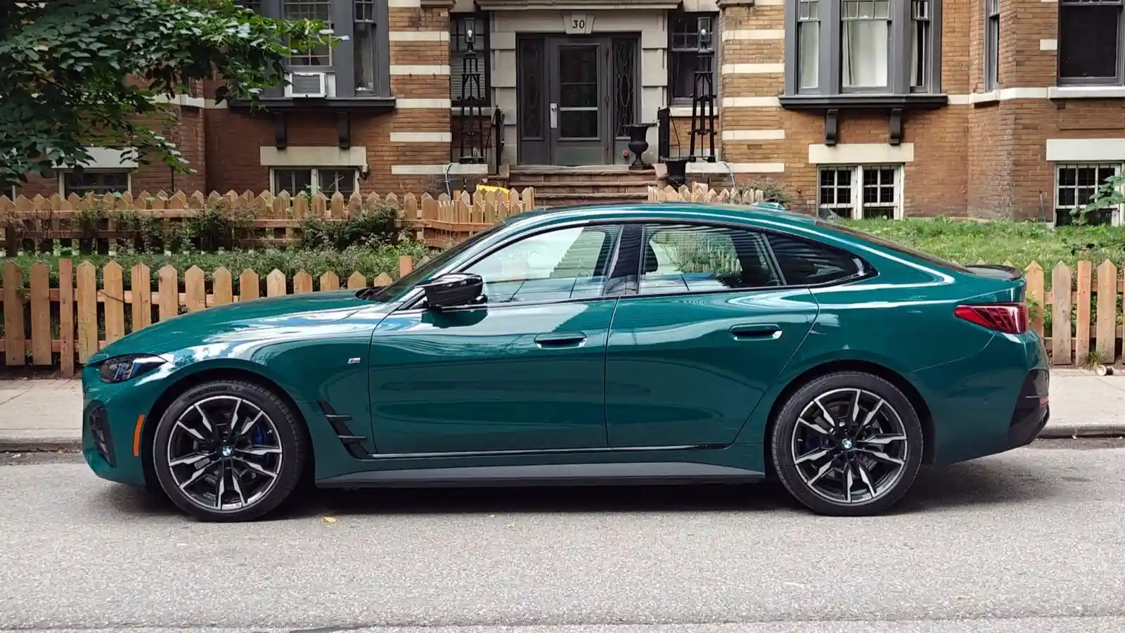 Perfil lateral de un BMW i4 M60 xDrive 2026 en color Cape York Green estacionado junto a la acera frente a un edificio de ladrillo con una valla de estacas