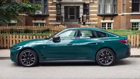 Perfil lateral de un BMW i4 M60 xDrive 2026 en color Cape York Green estacionado junto a la acera frente a un edificio de ladrillo con una valla de estacas