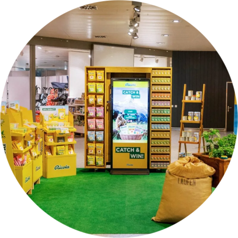 Ricola product display with yellow packaging, a green carpet, a digital sign showing 'Catch & Win', and a large sack labeled 'Laufen'.