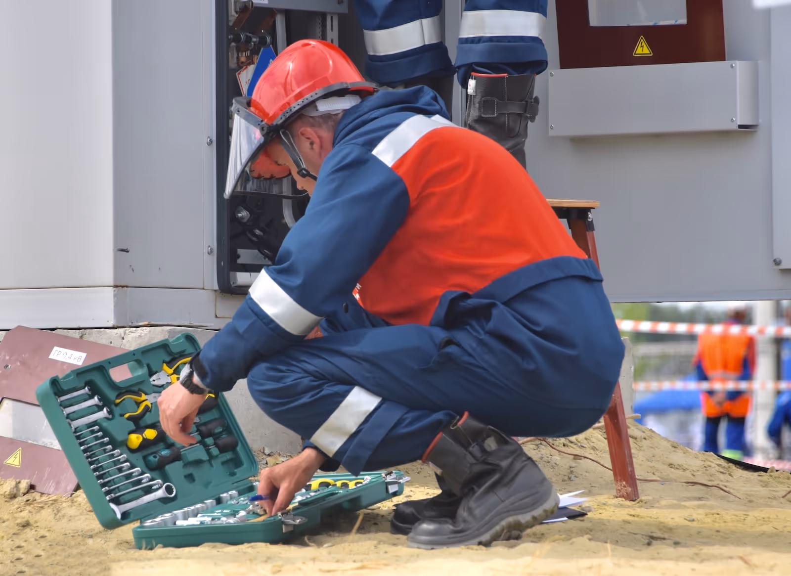 Electric technician in red and blue protective gear crouching and selecting tools from a green case near an open electrical panel outdoors.