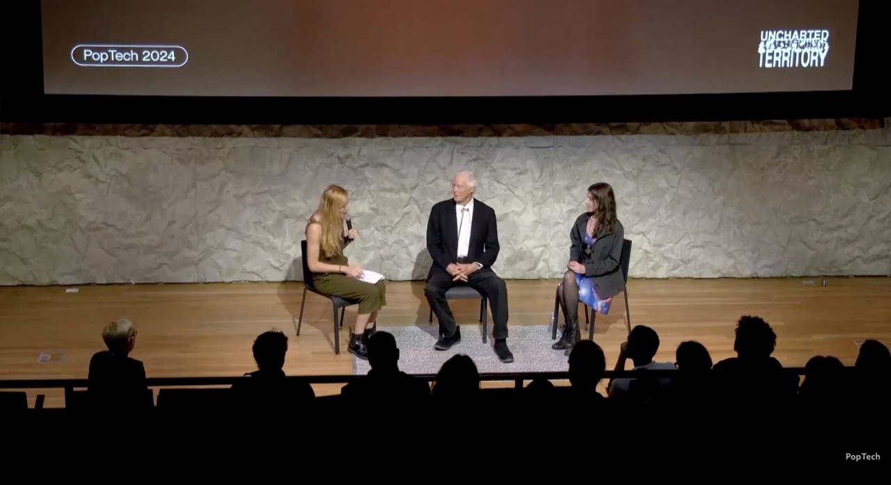 Three people seated on stage engaged in a discussion at PopTech 2024 with an audience in silhouette.