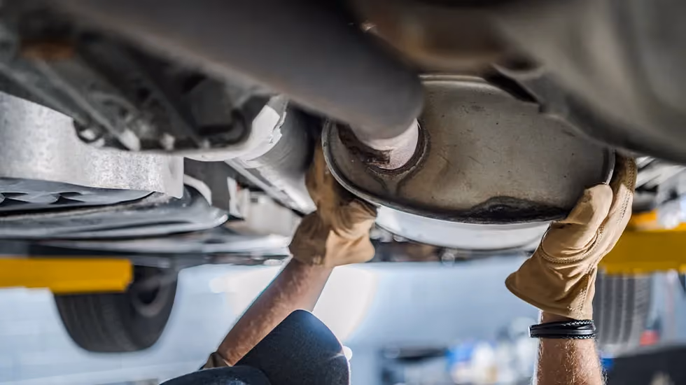In a repair shop, a car has been raised on a hydraulic lift. A mechanic is standing beneath it and is carefully handling the catalytic converter.