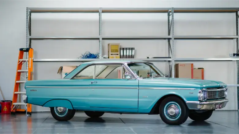 Classic turquoise vintage car parked indoors beside storage shelves and equipment.