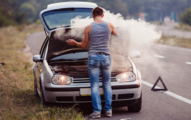 Stranded on the side of the road, a man stares at the smoke billowing out of his car’s engine. 