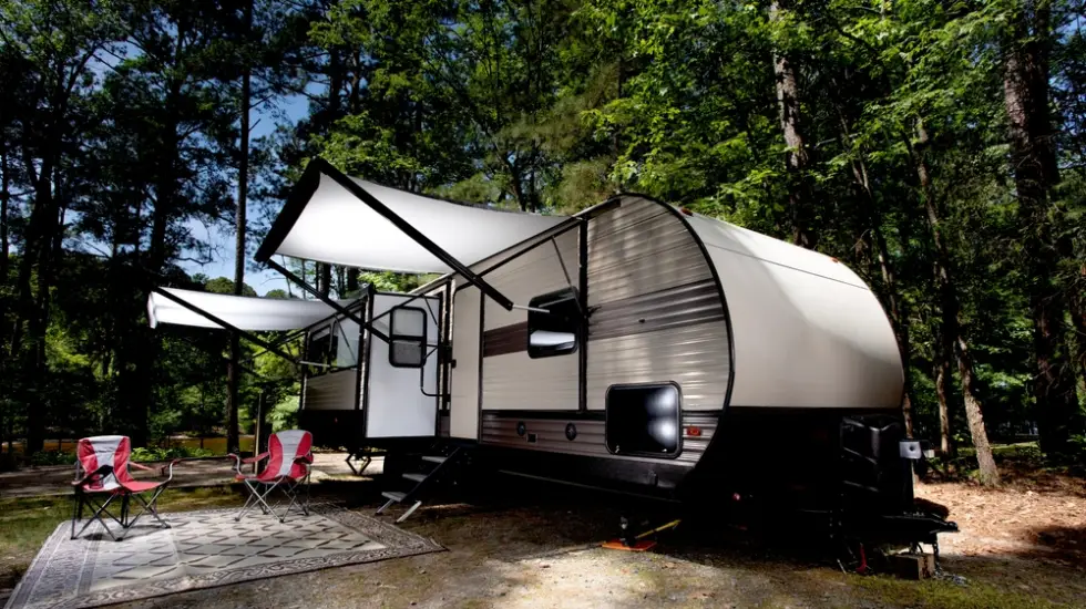 Smiling man handing over keys while an old white camper trailer is being towed away on a flatbed truck in a sunny outdoor setting.