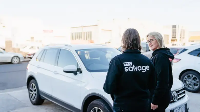 A 1800 Salvage worker in a black jacket talks with a woman standing beside a white Volkswagen SUV in a car park.