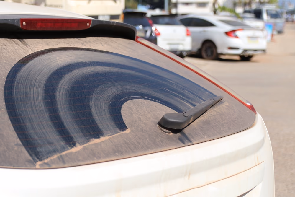 A close-up of a very dusty rear windscreen of a white car, showing a clean arched path cleared by the rear wiper blade.