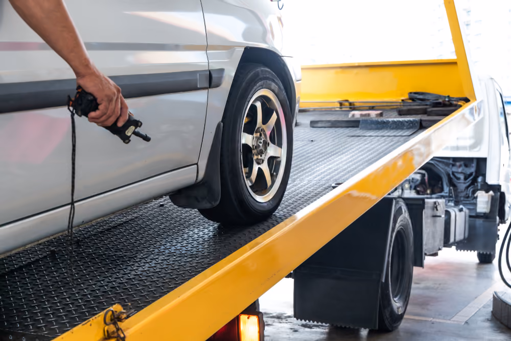 A close-up of a silver car being loaded onto a yellow tilt-tray tow truck as an operator uses a winch remote control.