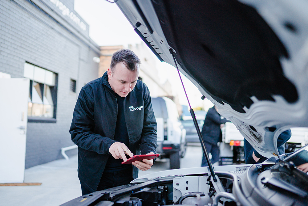 1800 Salvage team member inspecting a vehicle's engine using a tablet.