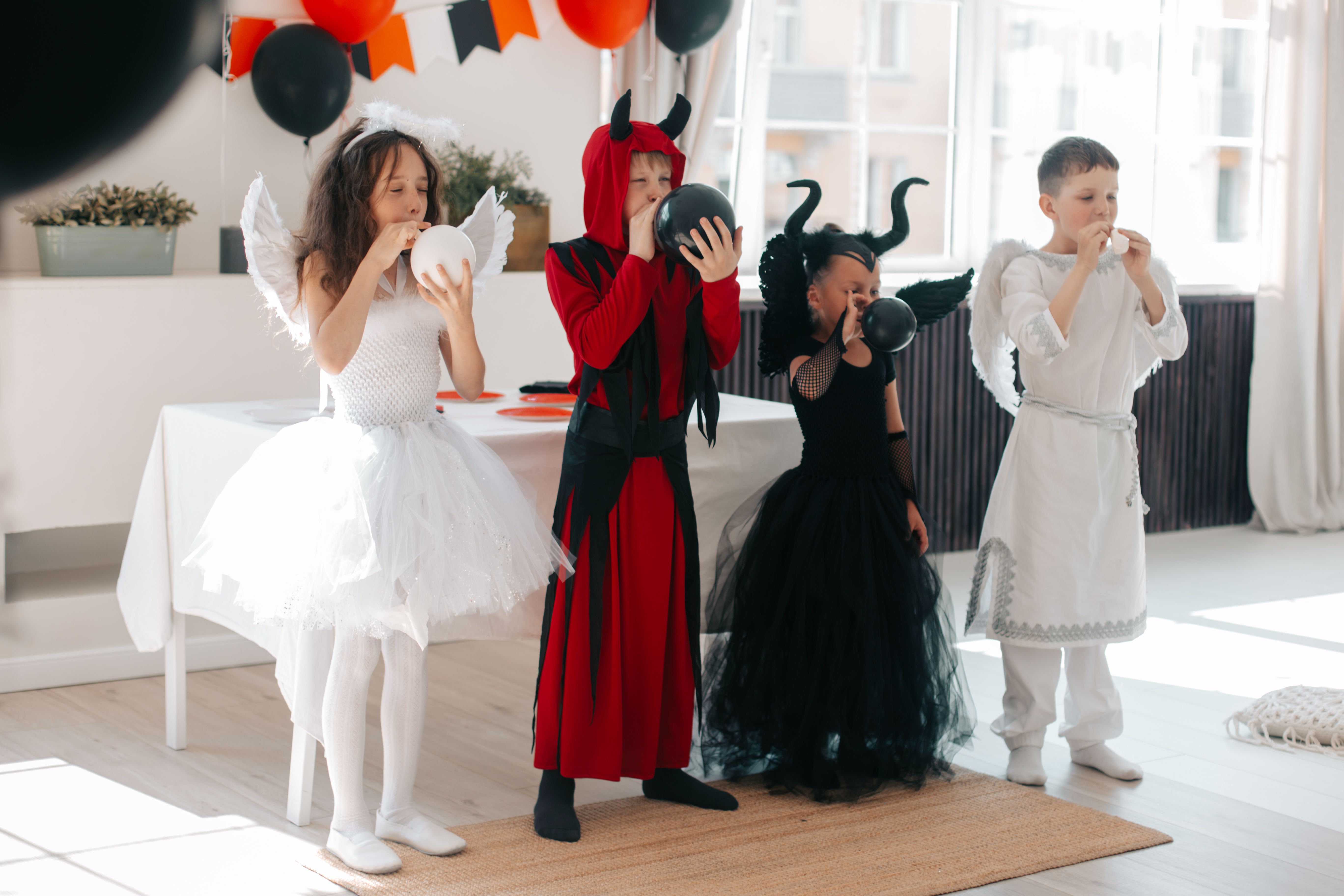 Children in different Halloween costumes in a line blowing up balloons at a party indoors.