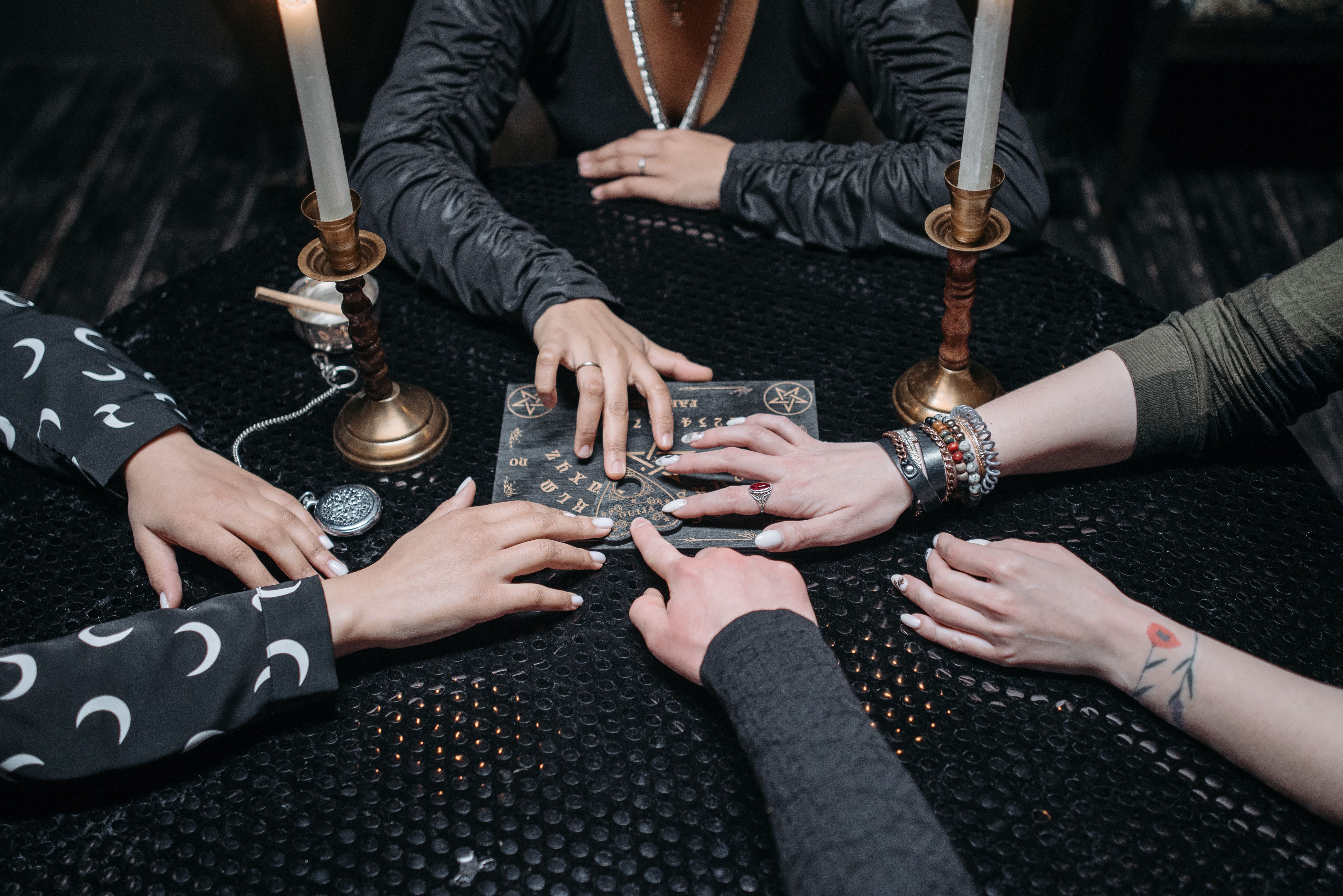 A group gathered around a Ouija board each with a finger on the board in a dimly lit room.