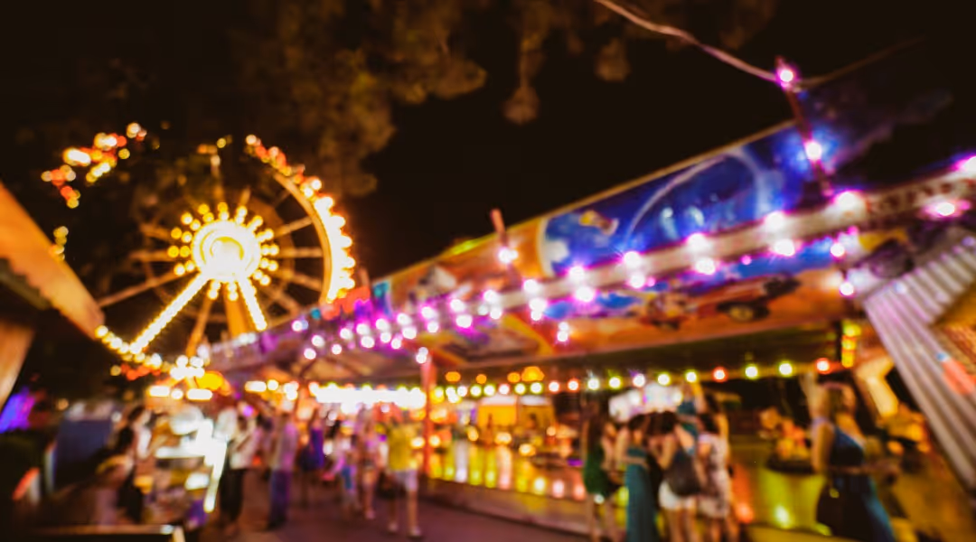Unscharfe Nachtaufnahme eines Jahrmarkts mit Menschen, hellen Lichtern und einem leuchtenden Riesenrad.