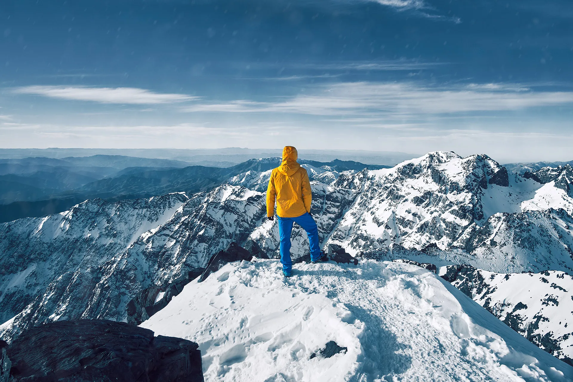 Mountaineer standing on a mountain peak overlooking snowy mountain range under a blue sky.