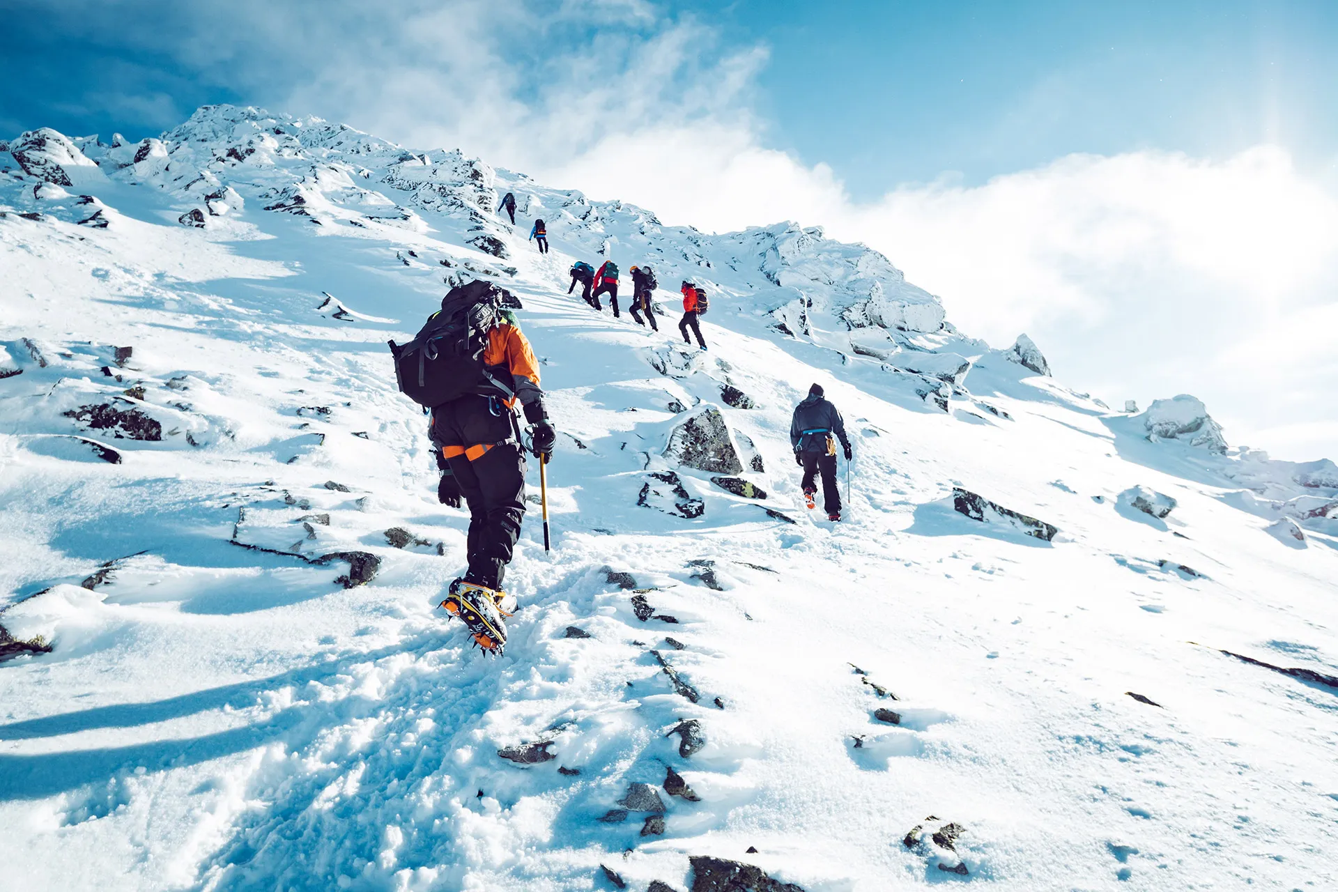 Group of mountain climbers ascending a snowy, rocky slope under a bright blue sky.