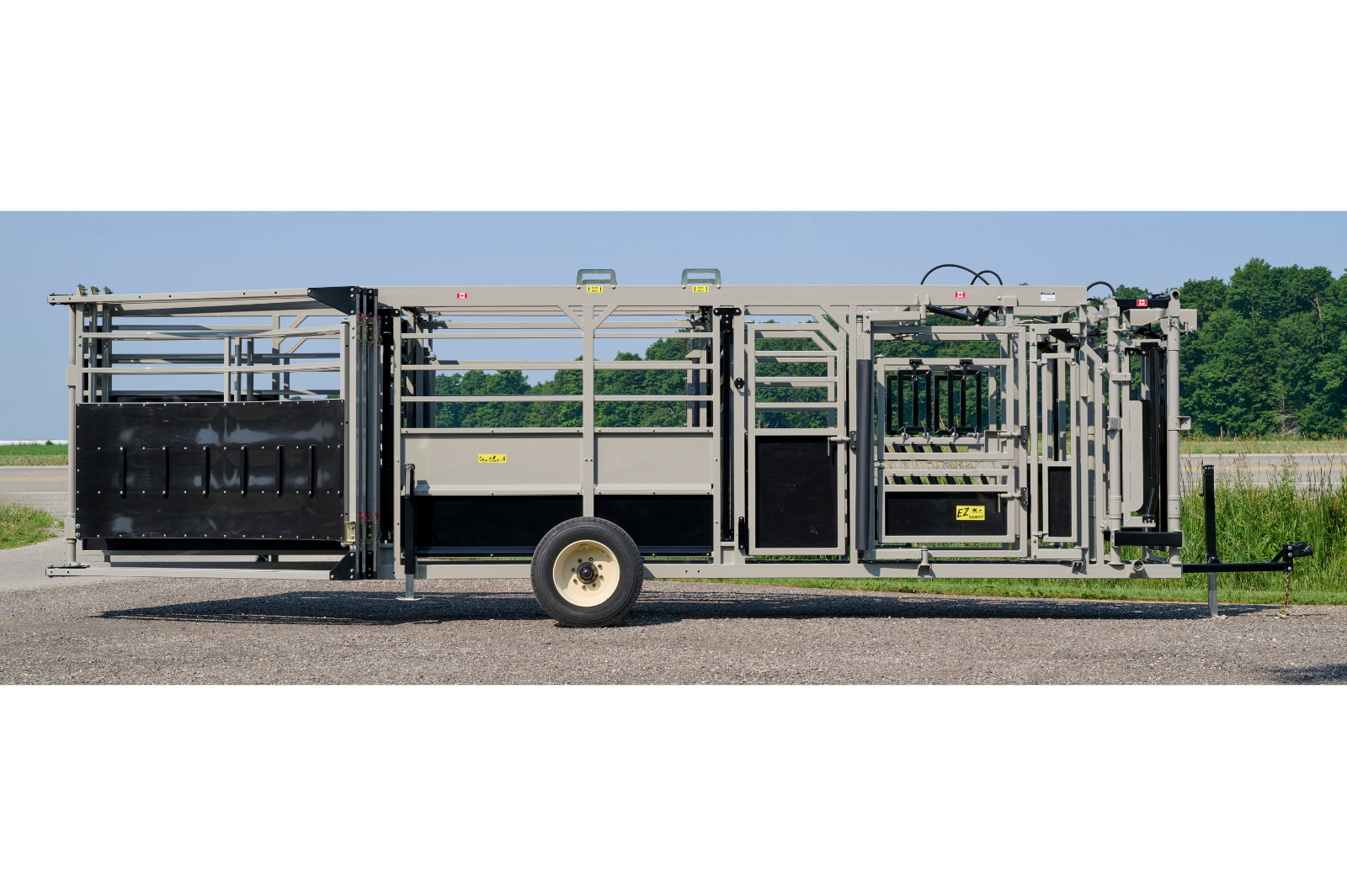 A livestock handling system with metal rails and gates, featuring wheels for mobility. The equipment is set up outdoors on a gravel surface, with grass and trees in the background.