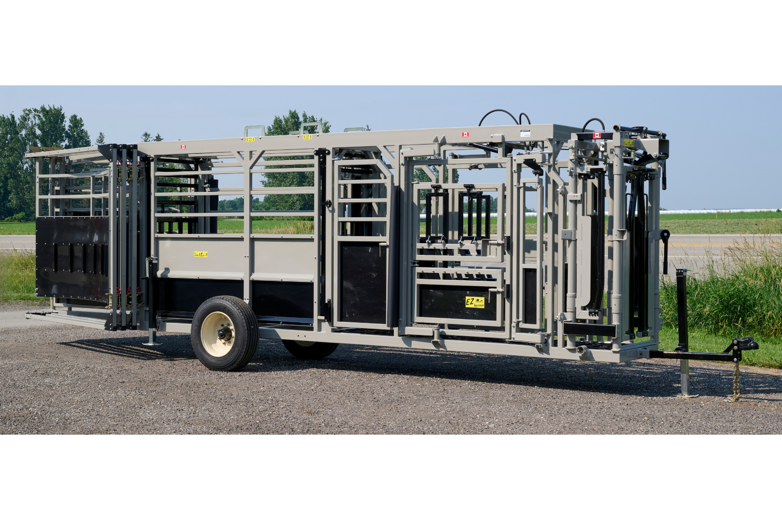 A livestock handling system with metal rails and gates, featuring wheels for mobility. The equipment is set up outdoors on a gravel surface, with grass and trees in the background.