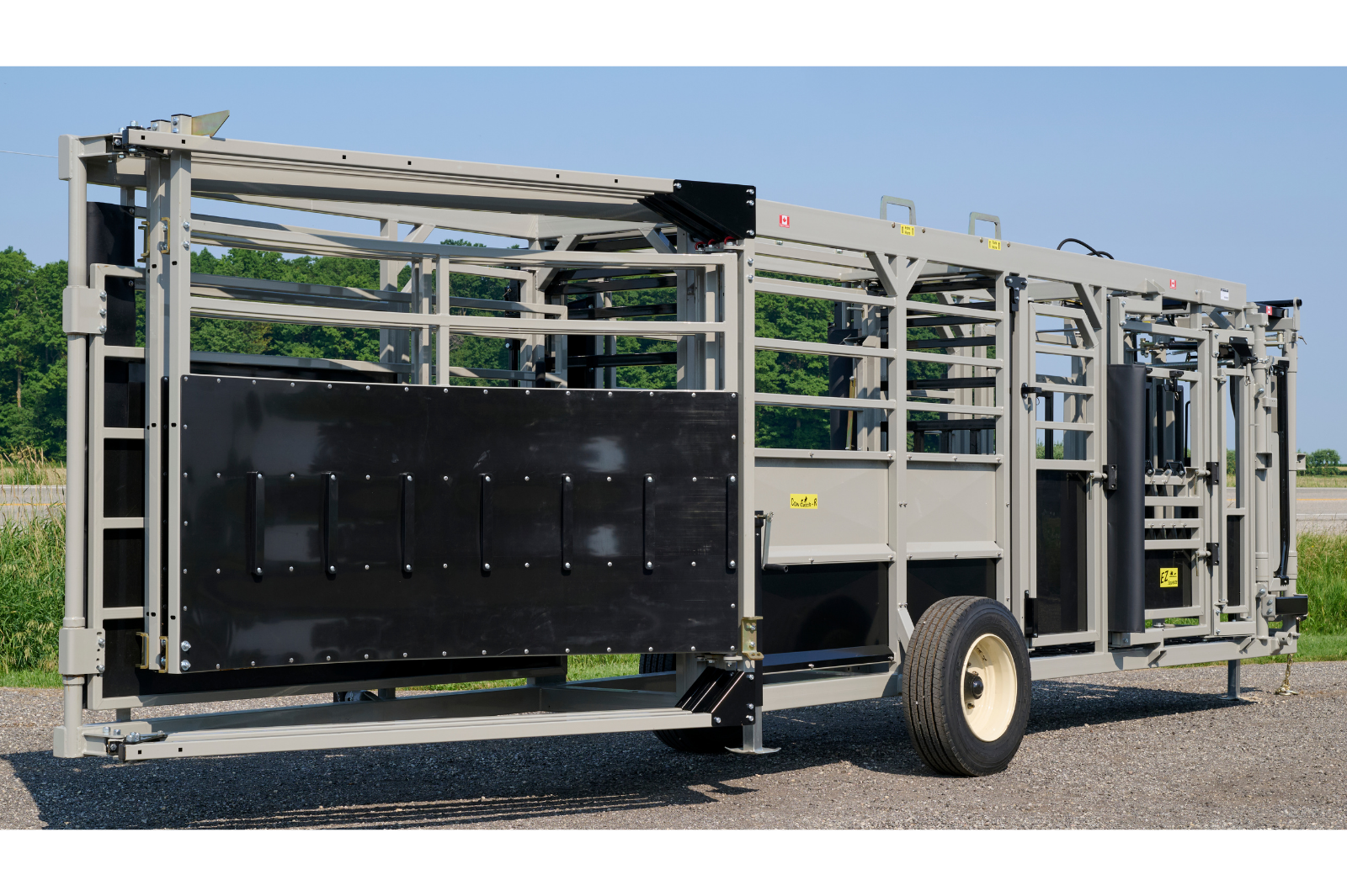 A livestock handling system with metal rails and gates, featuring wheels for mobility. The equipment is set up outdoors on a gravel surface, with grass and trees in the background.