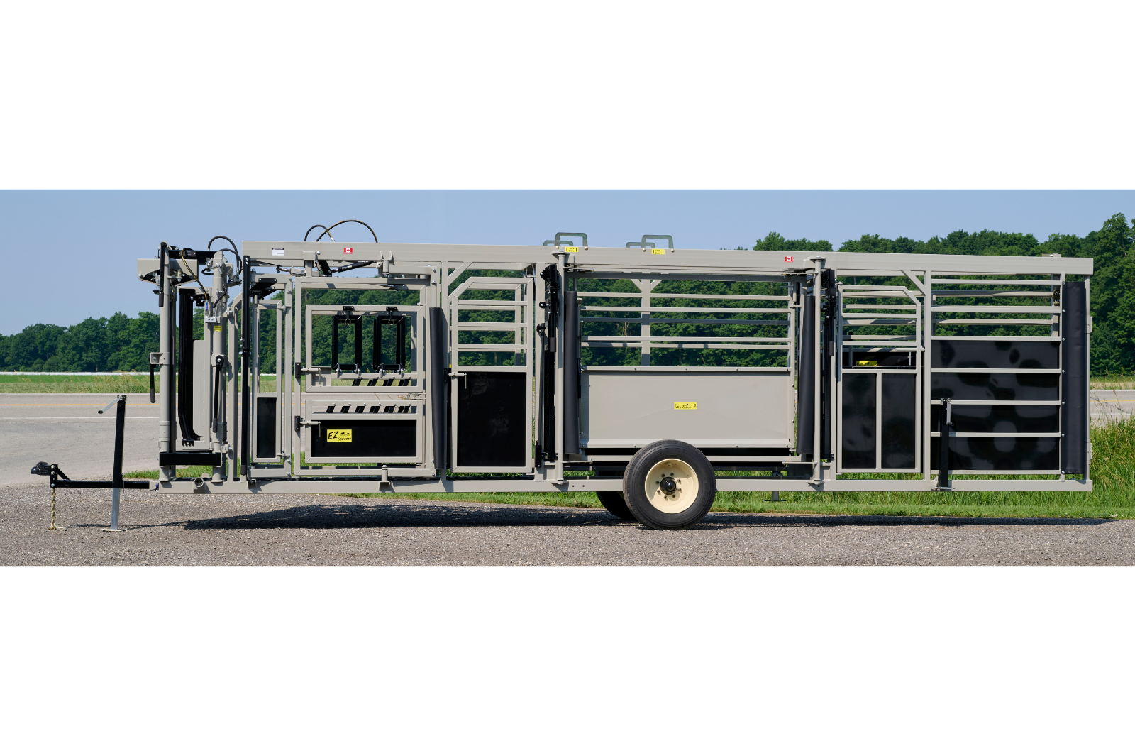 A livestock handling system with metal rails and gates, featuring wheels for mobility. The equipment is set up outdoors on a gravel surface, with grass and trees in the background.