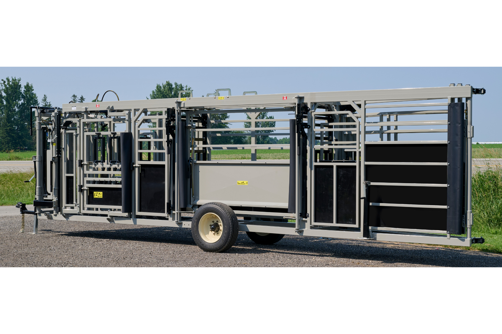 A livestock handling system with metal rails and gates, featuring wheels for mobility. The equipment is set up outdoors on a gravel surface, with grass and trees in the background.