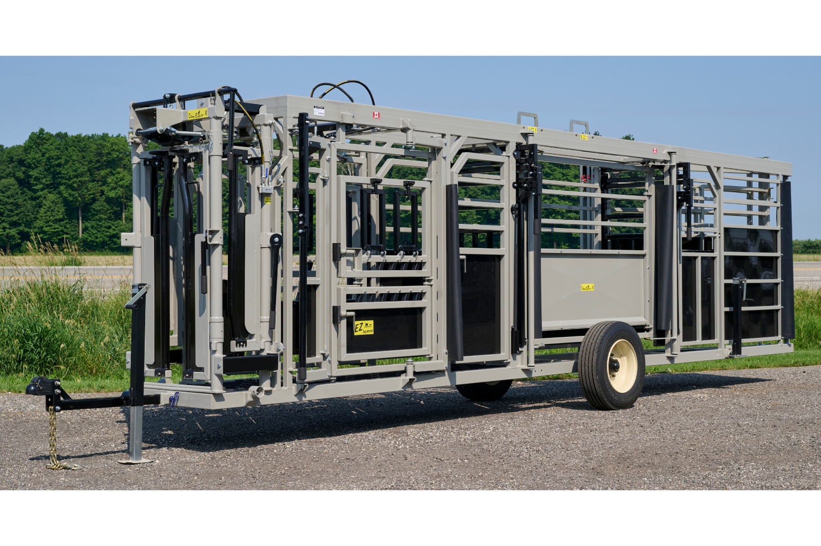 A livestock handling system with metal rails and gates, featuring wheels for mobility. The equipment is set up outdoors on a gravel surface, with grass and trees in the background.
