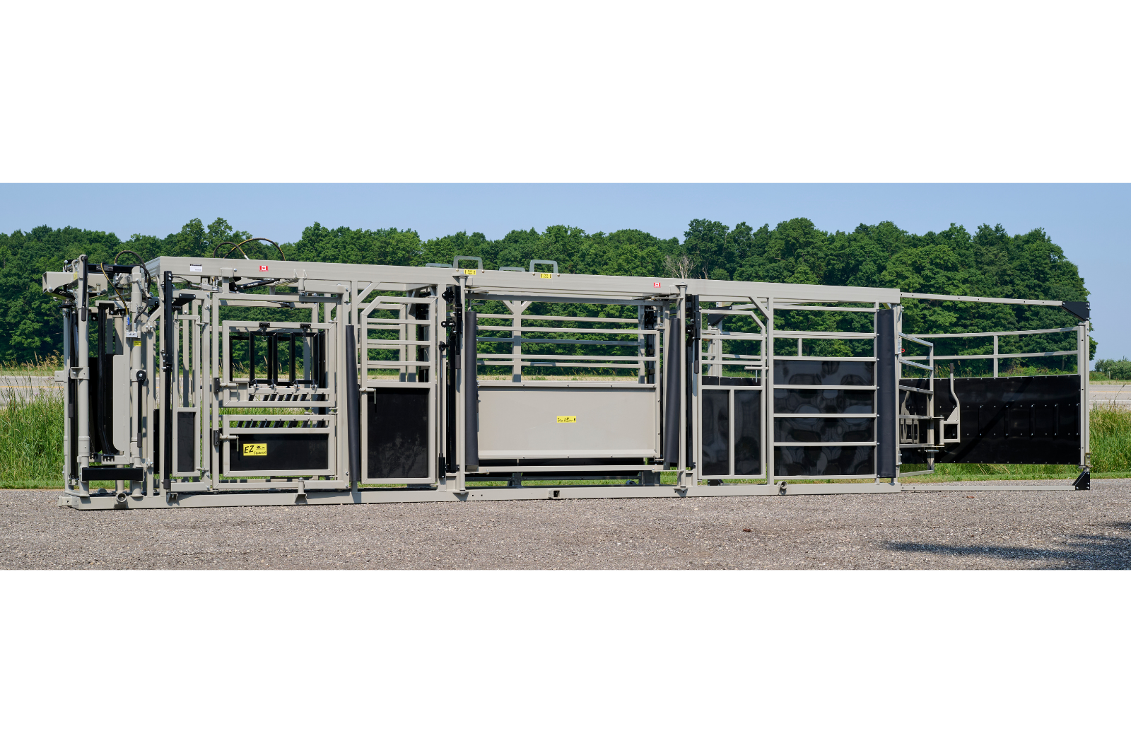 A livestock handling system with metal rails and gates, featuring wheels for mobility. The equipment is set up outdoors on a gravel surface, with grass and trees in the background.