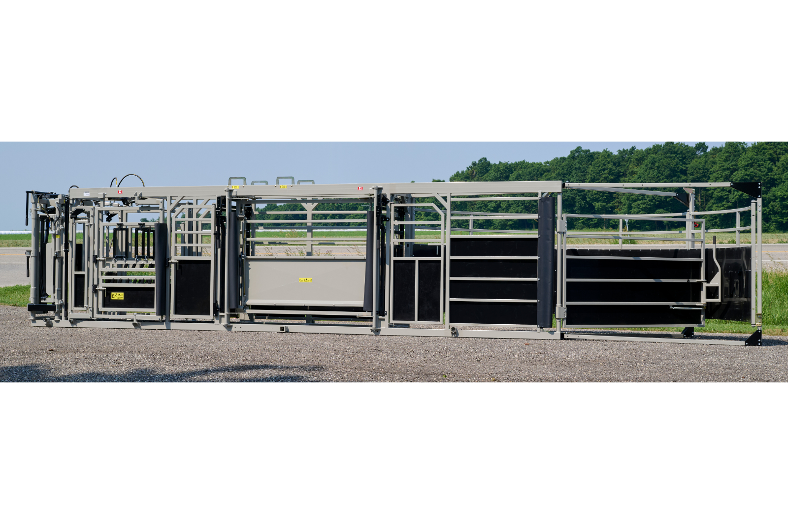 A livestock handling system with metal rails and gates, featuring wheels for mobility. The equipment is set up outdoors on a gravel surface, with grass and trees in the background.