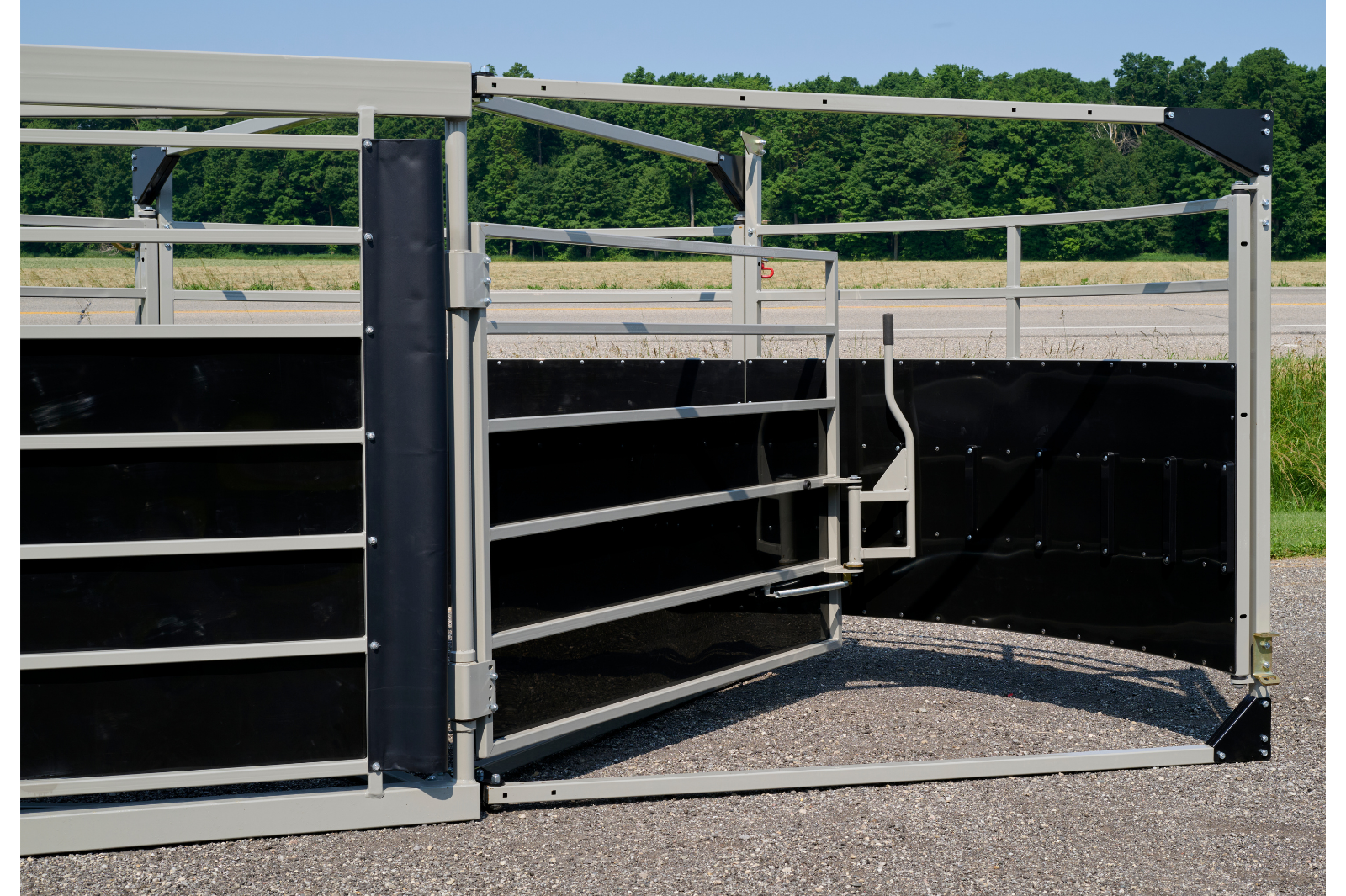 A livestock handling system with metal rails and gates, featuring wheels for mobility. The equipment is set up outdoors on a gravel surface, with grass and trees in the background.
