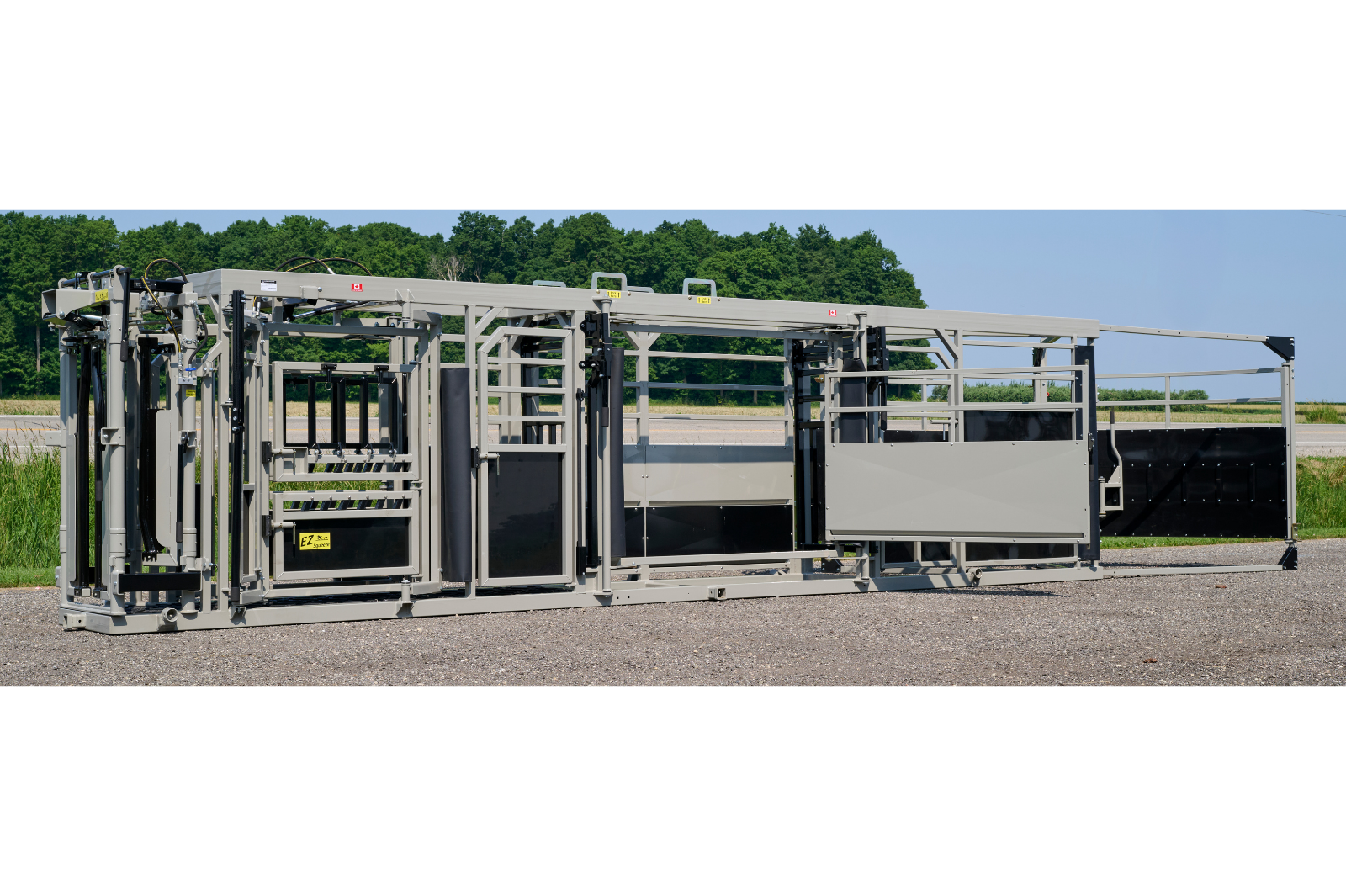 A livestock handling system with metal rails and gates, featuring wheels for mobility. The equipment is set up outdoors on a gravel surface, with grass and trees in the background.