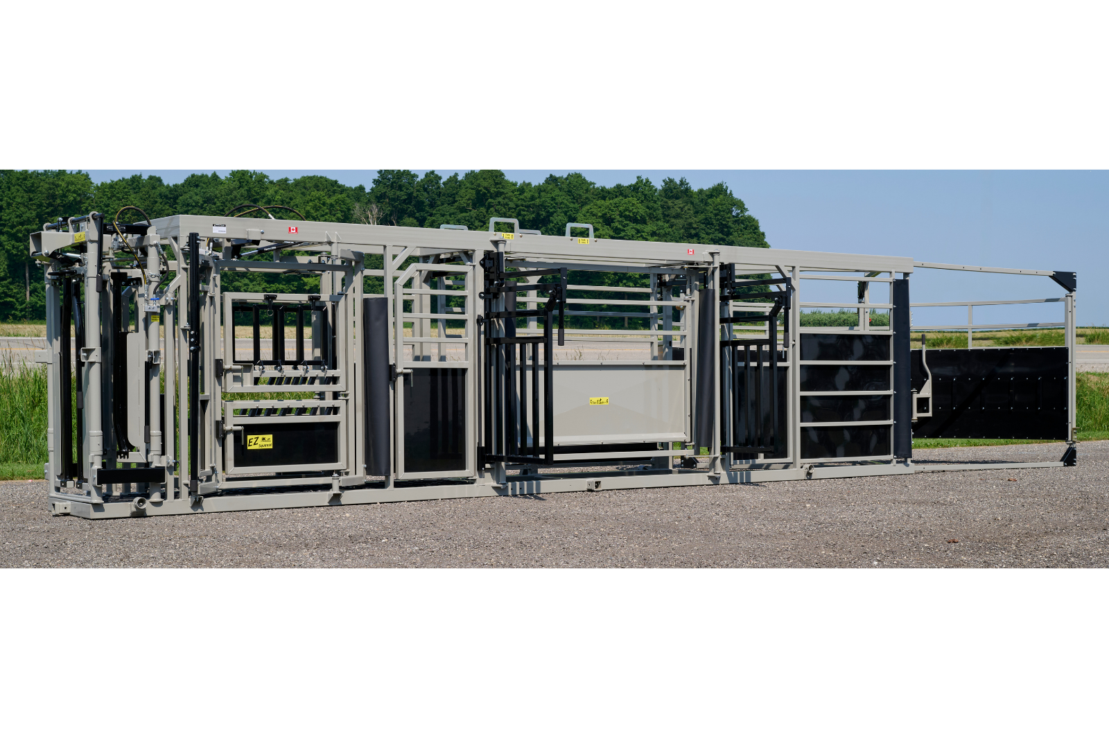 A livestock handling system with metal rails and gates, featuring wheels for mobility. The equipment is set up outdoors on a gravel surface, with grass and trees in the background.