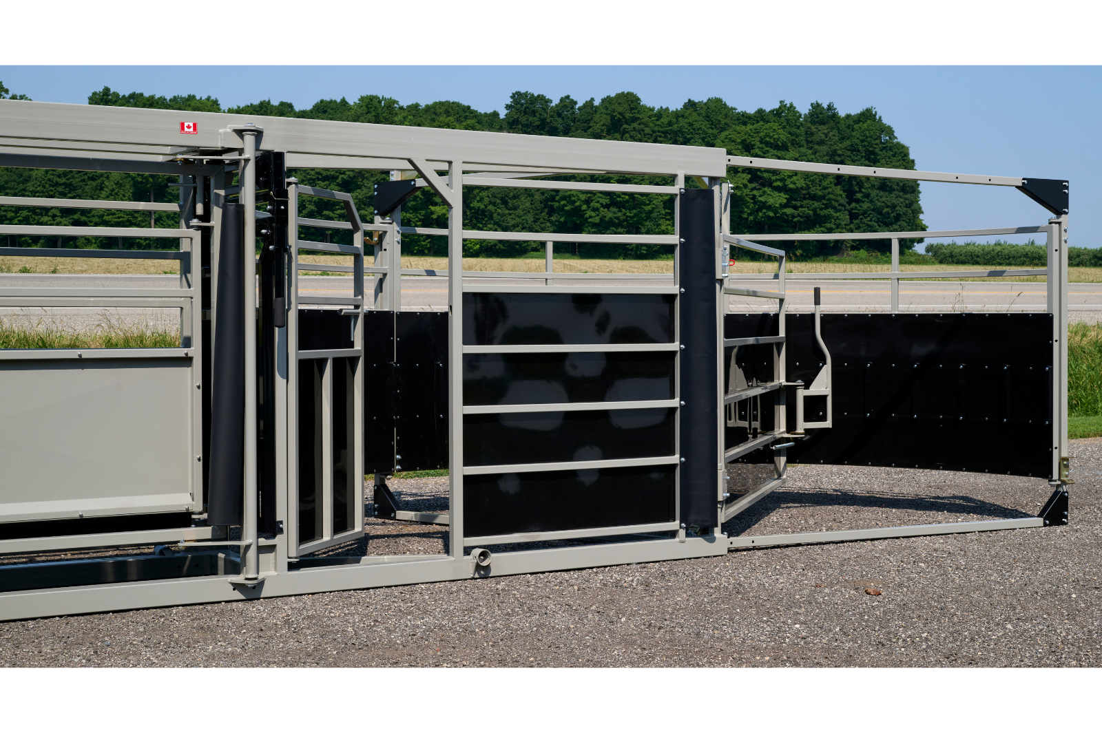 A livestock handling system with metal rails and gates, featuring wheels for mobility. The equipment is set up outdoors on a gravel surface, with grass and trees in the background.