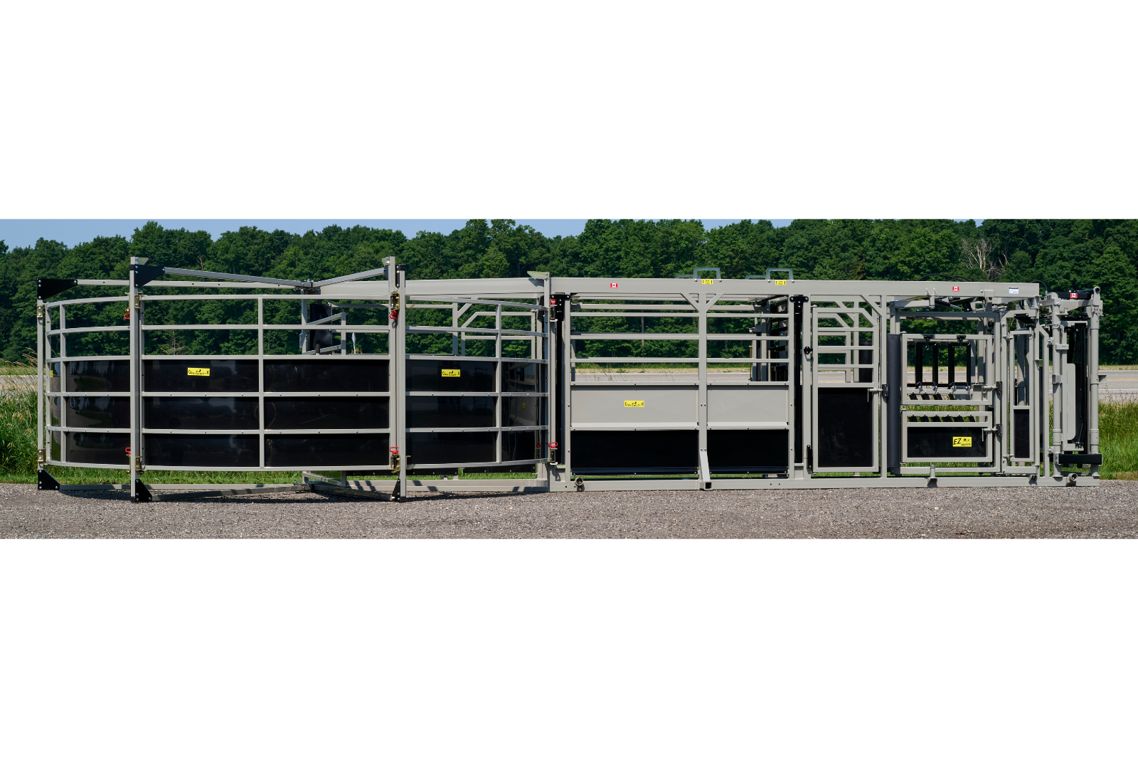 A livestock handling system with metal rails and gates, featuring wheels for mobility. The equipment is set up outdoors on a gravel surface, with grass and trees in the background.