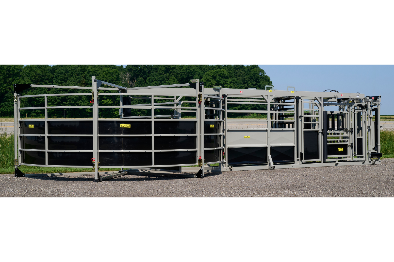 A livestock handling system with metal rails and gates, featuring wheels for mobility. The equipment is set up outdoors on a gravel surface, with grass and trees in the background.