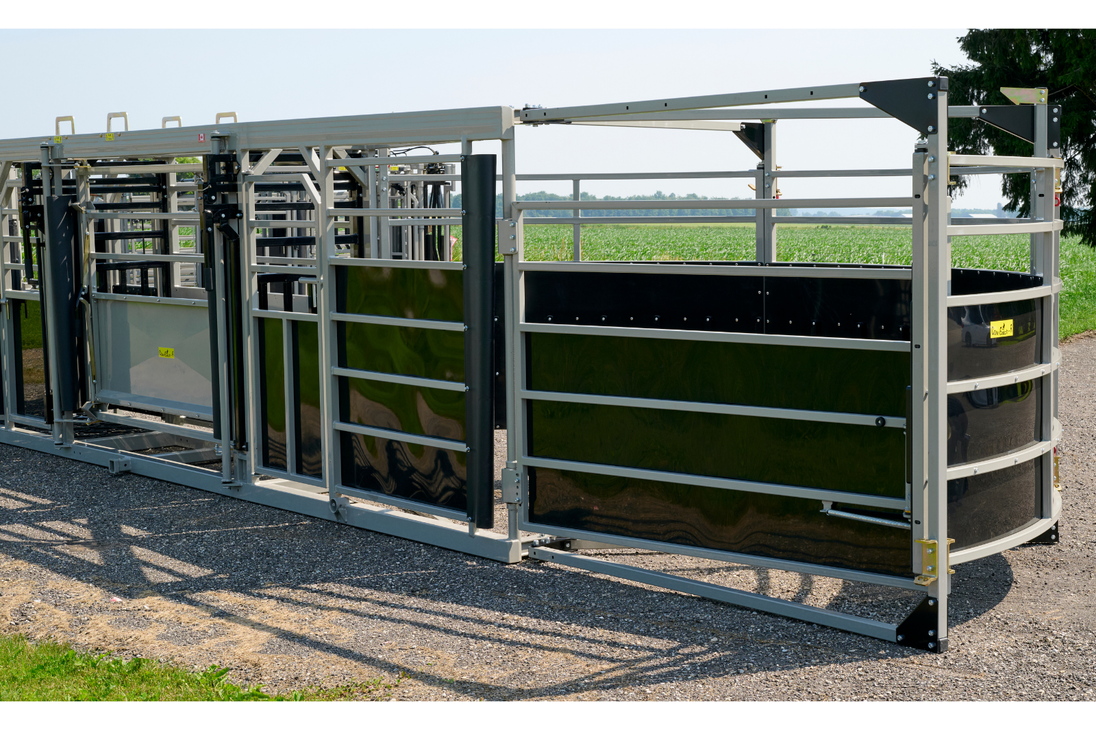 A livestock handling system with metal rails and gates, featuring wheels for mobility. The equipment is set up outdoors on a gravel surface, with grass and trees in the background.