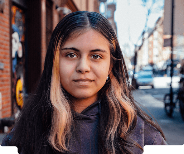 Photo of a young girl wearing a sweatshirt slightly smiling at the camera in an urban setting.