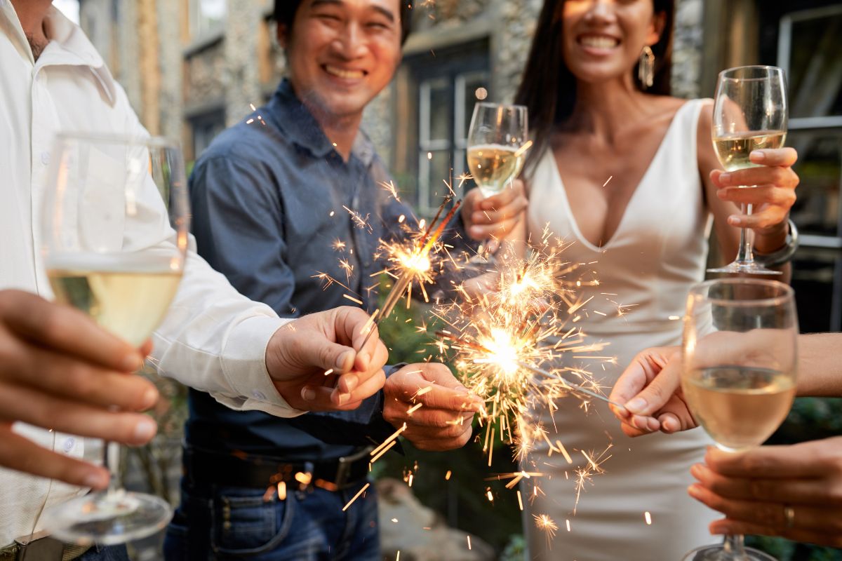 A group of people, dressed in formal attire and smiling, celebrate outdoors with sparklers and glasses of white wine.