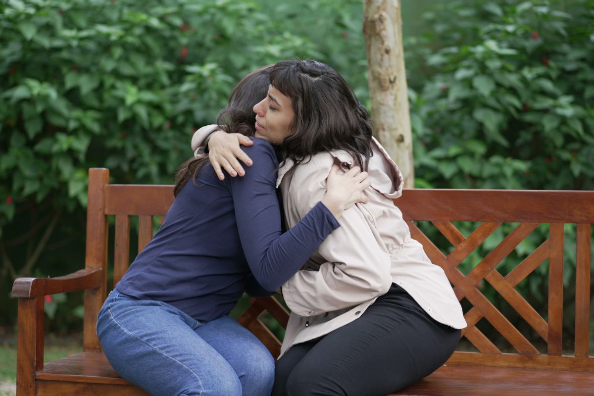Suffering woman being support by friend with an embrace while sitting outside on a park bench